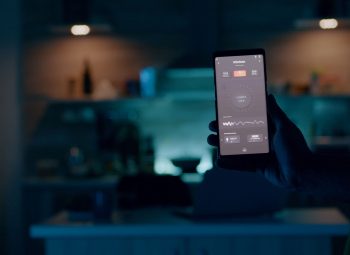 Person holding mobile phone with high tech application in smart house features controlling lights with wireless device. Man turning on the light sitting in home kitchen with automation lighting system